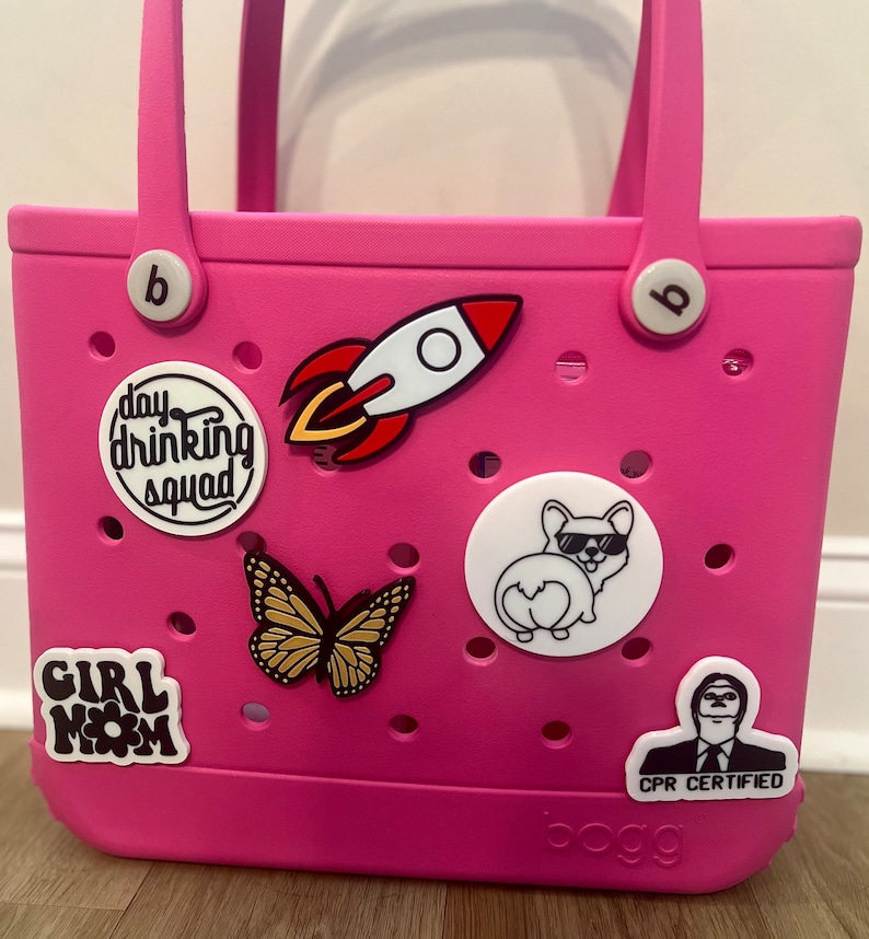 May include: Pink plastic tote bag with various colorful patches, including a rocket, a butterfly, a dog, and a person with the text "CPR Certified". The bag also has a white circle with the text "day drinking squad" and a white circle with the text "Girl Mom". The bag has two black handles and the word "BOGG" printed on the bottom.