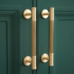 May include: Two gold cabinet pulls with a fluted design. The pulls are mounted on a dark green cabinet door.