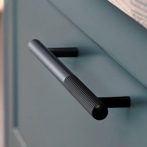 May include: Black cabinet door handle with a textured, ridged design. The handle is made of metal and has a rounded end.