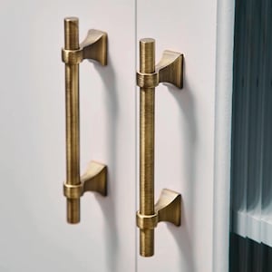 May include: Two gold cabinet pulls with a brushed finish and a simple, modern design. The pulls have a rectangular shape with rounded edges and a slightly curved bar.