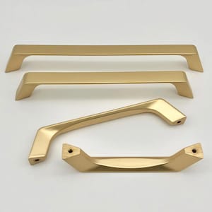 May include: Four gold-colored cabinet pulls in various shapes. Two are straight, one is curved, and one is a shorter, angled design. All have a matte finish and are set against a white background.