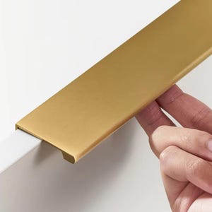 May include: A brushed gold cabinet pull with a rectangular shape and rounded edges. The pull is mounted on a white surface.
