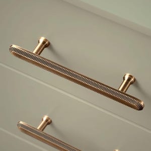 May include: Two brushed brass cabinet pulls with a textured, patterned surface. The pulls are rectangular in shape with rounded edges.