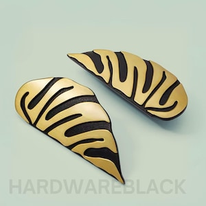 May include: Two gold and black leaf-shaped hair clips. The clips have a textured surface and a wavy pattern.