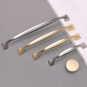May include: A collection of cabinet hardware, including silver and gold-toned handles and a round knob. The handles vary in size and feature a curved design. The hardware is displayed on a gray surface.