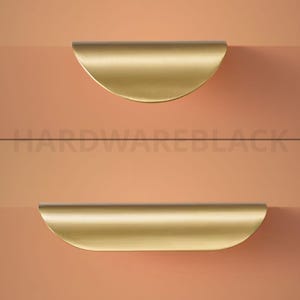 May include: Two gold-coloured, semi-circular cabinet pulls against a peach and coral background. The pulls are positioned horizontally, one above the other. The text "HARDWAREBLACK" is printed in black across the centre.
