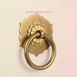 May include: A brass ring pull with a decorative floral design. The ring is attached to a brass plate with a scalloped edge.
