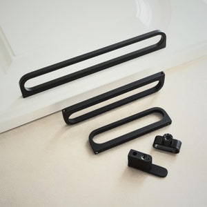 May include: Four black metal cabinet pulls with a rectangular shape and a cutout in the centre. The pulls are in different sizes.
