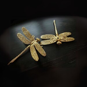 Puede incluir: Dos tiradores de caj&oacute;n de lib&eacute;lula de color dorado con detalles intrincados. Los tiradores de caj&oacute;n est&aacute;n sobre una superficie de madera oscura.