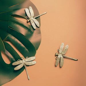 Puede incluir: Tres broches de lib&eacute;lula plateados sobre un fondo de hoja verde. La hoja est&aacute; sobre una superficie marr&oacute;n claro.