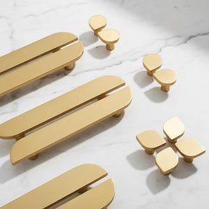 May include: A collection of gold-colored cabinet hardware, including long, rectangular pulls with a split down the middle and various knob designs. The hardware is arranged on a white marble surface, showcasing a modern aesthetic.