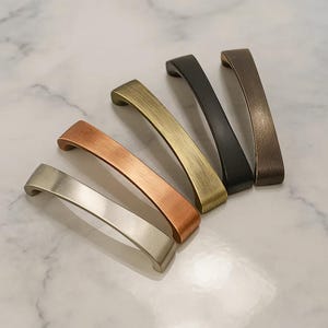 May include: A collection of five cabinet pulls in various metallic finishes: silver, copper, brass, black, and bronze. Each pull has a curved, rectangular design, perfect for updating kitchen or bathroom cabinetry. The pulls are arranged on a marble surface.