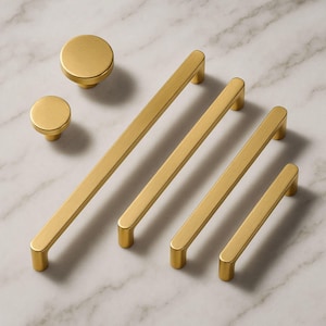May include: A collection of gold-colored cabinet hardware, including two knobs and four bar pulls of varying lengths. The hardware has a brushed finish and a modern, minimalist design. The hardware is displayed on a white and gray marble surface.
