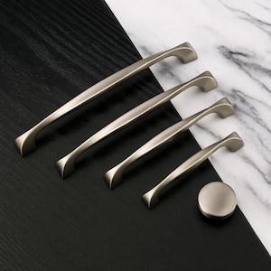 May include: A collection of four brushed nickel cabinet pulls and a matching knob displayed on a black and white marble surface. The pulls are of varying sizes, featuring a curved handle design. The knob is round and has a similar finish.