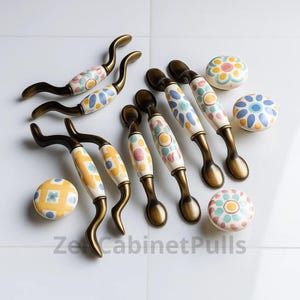 May include: A selection of cabinet handles and knobs. The handles feature a curved, bronze-coloured metal base with a white ceramic centre. The ceramic is decorated with colourful floral and polka dot patterns. The knobs have a similar design.