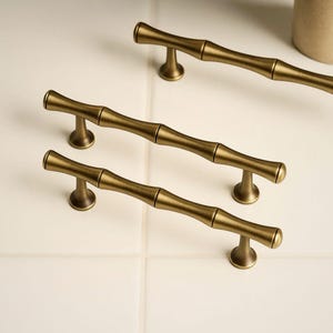 May include: Four antique brass cabinet pulls with a bamboo design. The pulls are a warm, golden color and have a textured surface. Each pull is approximately 6 inches long.