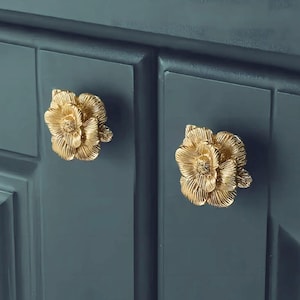 May include: Two gold-toned flower-shaped cabinet knobs with a detailed design. The knobs are mounted on a dark blue cabinet.