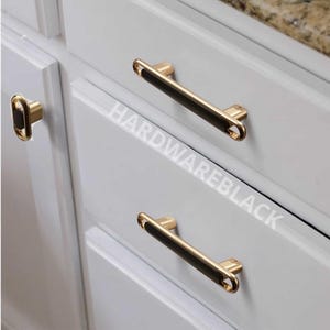 May include: Cabinet hardware with a gold finish and black accents. The image shows a close-up of cabinet doors with matching handles and a knob. The text "HARDWAREBLACK" is visible on the cabinet door.