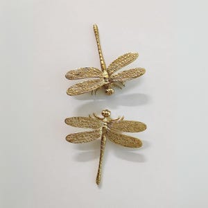 Puede incluir: Dos figuritas de lib&eacute;lula de metal dorado. Las lib&eacute;lulas tienen alas y cuerpos detallados.