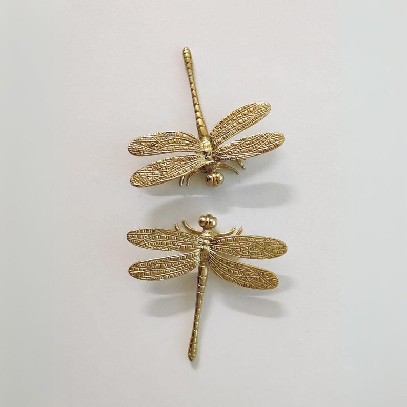 Puede incluir: Dos figuritas de lib&eacute;lula de metal dorado. Las lib&eacute;lulas tienen alas y cuerpos detallados.