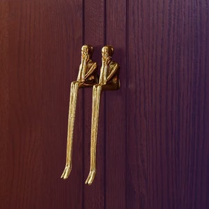 May include: Two gold-toned decorative door handles shaped like figures with their hands covering their faces. The figures are sitting with their legs extended.