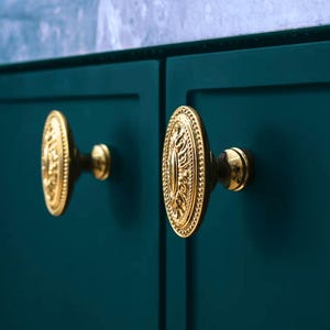 May include: Two gold oval cabinet knobs with ornate designs on a teal green cabinet door.
