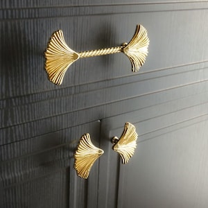 May include: Two gold-toned cabinet pulls with a fan-shaped design. The larger pull has a twisted bar connecting the two fan shapes. The smaller pull is a single fan shape.