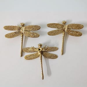 Puede incluir: Tres tiradores de caj&oacute;n de lib&eacute;lula de color dorado con detalles intrincados. Cada tirador tiene un agujero en el centro para el montaje.