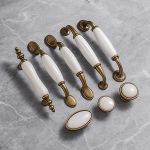 May include: A selection of cabinet hardware with white ceramic handles and antique brass-toned metal details. The set includes different handle styles and knob designs, suitable for furniture or cabinetry. The hardware is displayed on a grey marble surface.