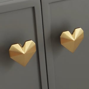 May include: Two gold heart-shaped cabinet knobs with a geometric design. The knobs are mounted on a gray cabinet door.