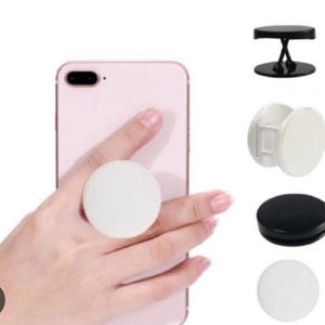 May include: A white round phone grip attached to a pink smartphone. Additional phone grips in black and white are displayed. The phone grip is a circular accessory that adheres to the back of a phone for a secure hold.