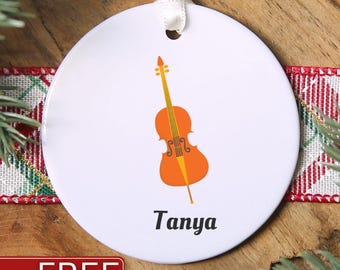 May include: A round white ceramic ornament featuring an orange cello illustration centered above the name Tanya in black text. The ornament includes a small hole at the top with a white ribbon loop for hanging.