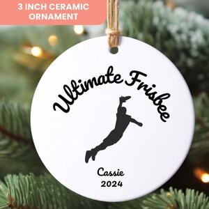 May include: White ceramic ornament with a black silhouette of a person playing ultimate frisbee. The text "Ultimate Frisbee" is written in a black cursive font above the silhouette. The text "Cassie 2024" is written in black below the silhouette.