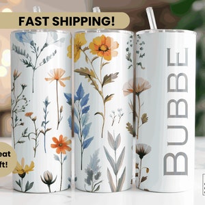 May include: Three stainless steel tumblers with a floral design. The tumbler on the left has a white background with a floral pattern in shades of blue, yellow, and orange. The tumbler in the middle has a white background with a floral pattern in shades of blue, yellow, and orange. The tumbler on the right has a white background with the word "BUBBE" written in gray letters.