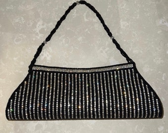 Puede incluir: Bolso de mano negro con cuentas con filas verticales de brillantes pedrería. El borde superior está forrado con una fila de pedrería. El asa es un cordón negro retorcido. Un elegante bolso de noche.