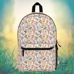 May include: A white backpack with a floral pattern featuring pink, yellow, and blue watercolor flowers and leaves. The backpack has a black zipper and a black adjustable strap.