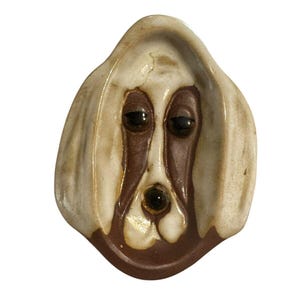 May include: A ceramic dish shaped like a dog face with long, drooping ears. The piece features a cream-colored glaze on the ears and forehead, with a dark brown glaze defining the eyes, nose, and muzzle area. The dog has a sad expression.