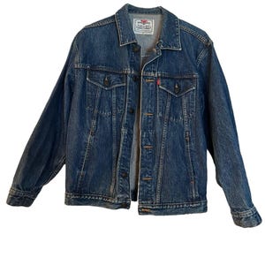 May include: A medium-wash blue denim jacket featuring a classic collar, long sleeves with buttoned cuffs, and a front button closure. The garment includes two chest pockets with buttoned flaps, a small red brand tab, and a Levi's label at the neck.