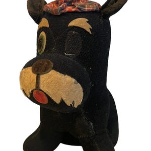 May include: A vintage black plush dog toy with a winking eye and a plaid hat. The dog has tan accents on its snout and ears, and a red tongue. The toy is a classic example of a stuffed animal.