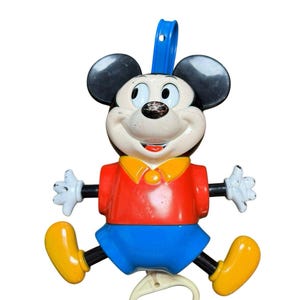 May include: A vintage plastic pull-string toy featuring a Mickey Mouse character with a black head, large ears, a red shirt with a yellow collar, blue shorts, white gloves, and yellow shoes. A blue hanging loop is at the top and a white ring at the bottom.