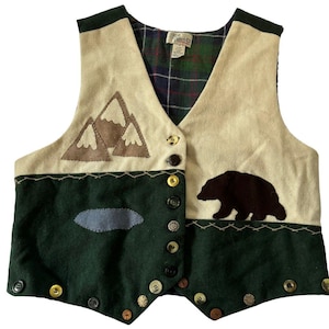 May include: A sleeveless vest featuring a cream-colored upper section with brown mountain appliqués and a dark green lower section with a blue pond appliqué. A dark brown bear silhouette is on the right. It has a plaid lining and mismatched buttons.