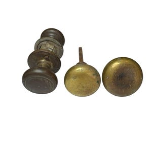 May include: Three door knobs are displayed. One is a dark brown, round knob with a metal mounting plate and internal mechanism. The other two are round, brass-colored knobs with a weathered, tarnished finish, each featuring a central metal spindle.