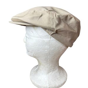 May include: A solid beige flat cap featuring a structured crown and a short, stiff brim. The hat is constructed from a woven fabric with visible stitching details along the side panels, providing a clean and uniform appearance throughout the design.
