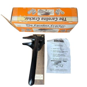 May include: A black metal nutcracker mounted on a rectangular wooden base, accompanied by its original orange cardboard box labeled The Carolina Cracker and a white paper instruction sheet detailing the four-step nut cracking process.