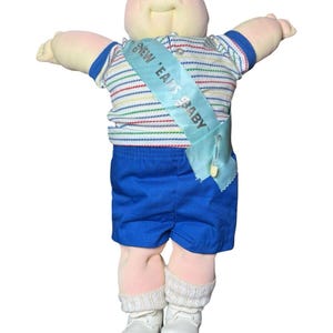 May include: A soft-sculpture doll with a round face and green eyes, wearing a white shirt with thin multicolored stripes, blue shorts, white socks, and white shoes. A light blue sash across the chest features the text NEW EARS BABY.