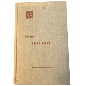 May include: A vintage book titled "Proces" by Franz Kafka. The book has a light tan linen cover with the title and author's name in a reddish-brown font. The cover also features the Roman numerals "XX" in a square.