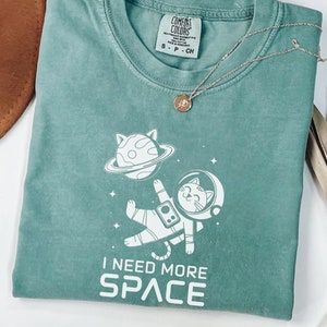 May include: A teal blue t-shirt with a white graphic of a cat in a spacesuit holding a planet. The text "I need more space" is printed below the graphic.