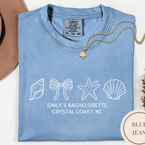 May include: A pale blue t-shirt featuring white illustrations of a seashell, bow, starfish, and shell. The text reads "EMILY'S BACHELORETTE CRYSTAL COAST, NC 2026". A gold necklace with a star pendant rests on the shirt.