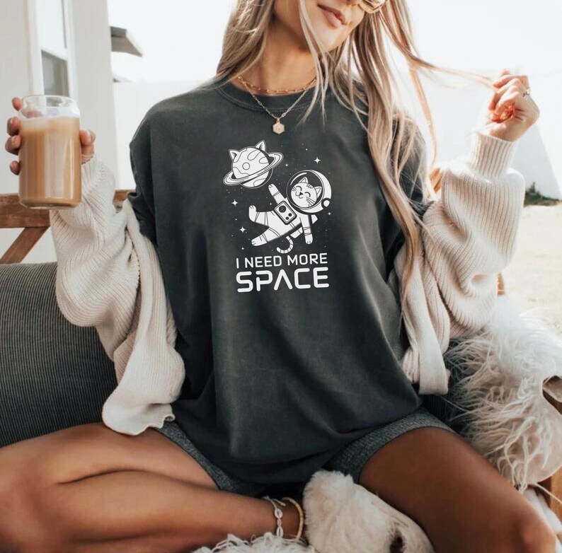 May include: A black sweatshirt with a white graphic of a cat in a spacesuit floating in space. The text "I need more space" is printed below the graphic.
