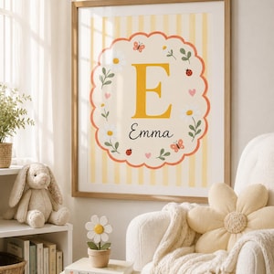 Könnte beinhalten: Ein gerahmter Wandkunstdruck mit einem großen gelben Buchstaben E und dem Namen Emma in schwarzer Schreibschrift. Das Design umfasst einen gewellten Rand, weiße Gänseblümchen, kleine rosa Herzen, Marienkäfer und Schmetterlinge auf gelb gestreiftem Grund.
